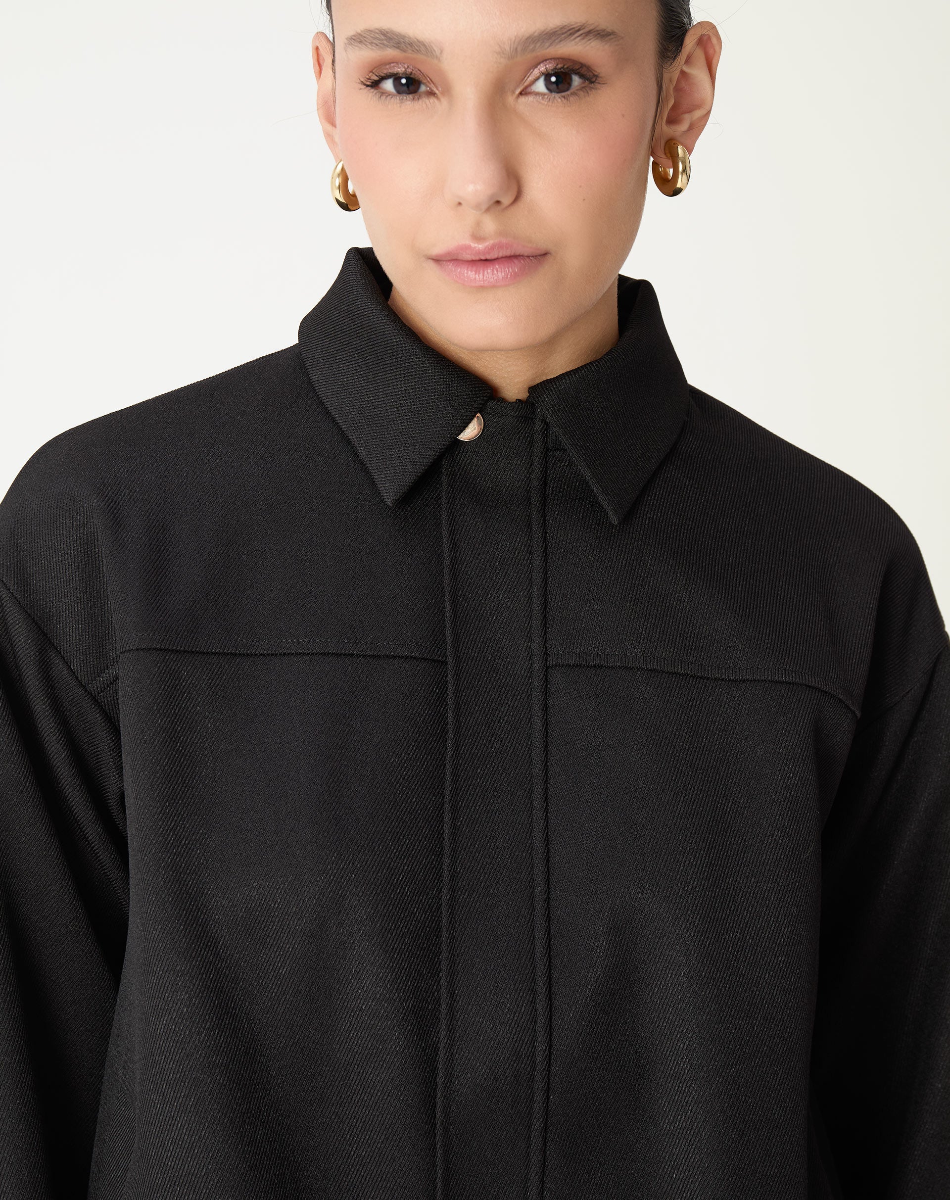 Detalhe frontal do casaco preto mostrando costura tipo pala e fechamento central com placket; tecido plano e acabamento clean