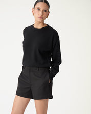 Mulher usando shorts alfaiataria preto cintura média com suéter preto, vista frontal com mãos nos bolsos, look casual-chic