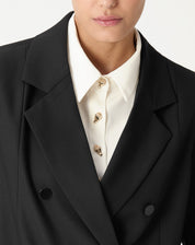 Detalhe do decote: camisa branca com botões dourados sob o blazer preto, ressaltando lapela e acabamento do pescoço.