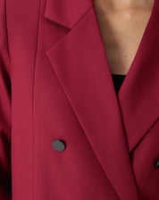 Detalhe frontal com as mãos da modelo nos bolsos embutidos do blazer vinho, mostrando o caimento solto.