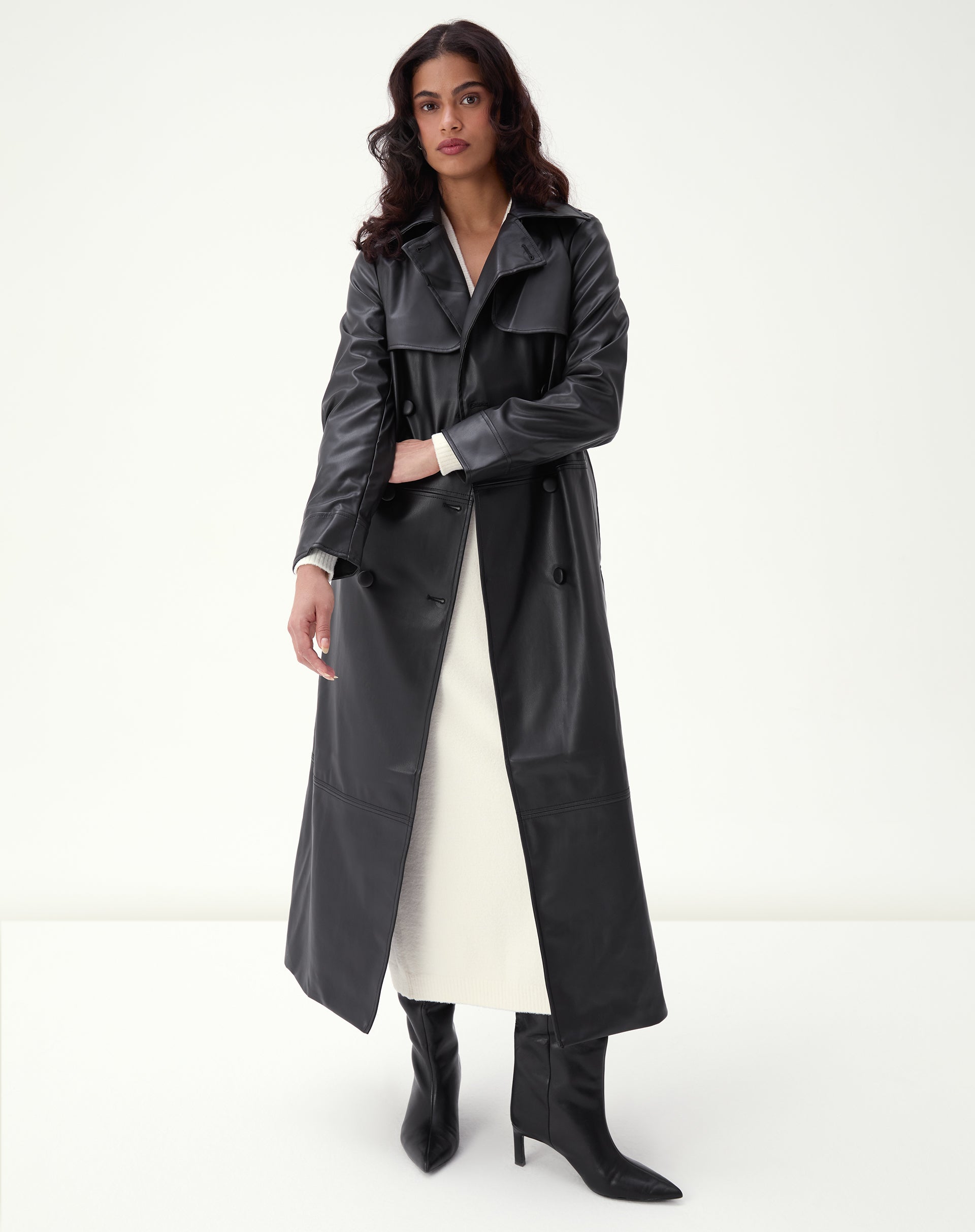 Modelo vestindo trench coat longo PU preto com cinto amarrado, abotoamento duplo e botas pretas em estúdio de fundo branco, look completo.