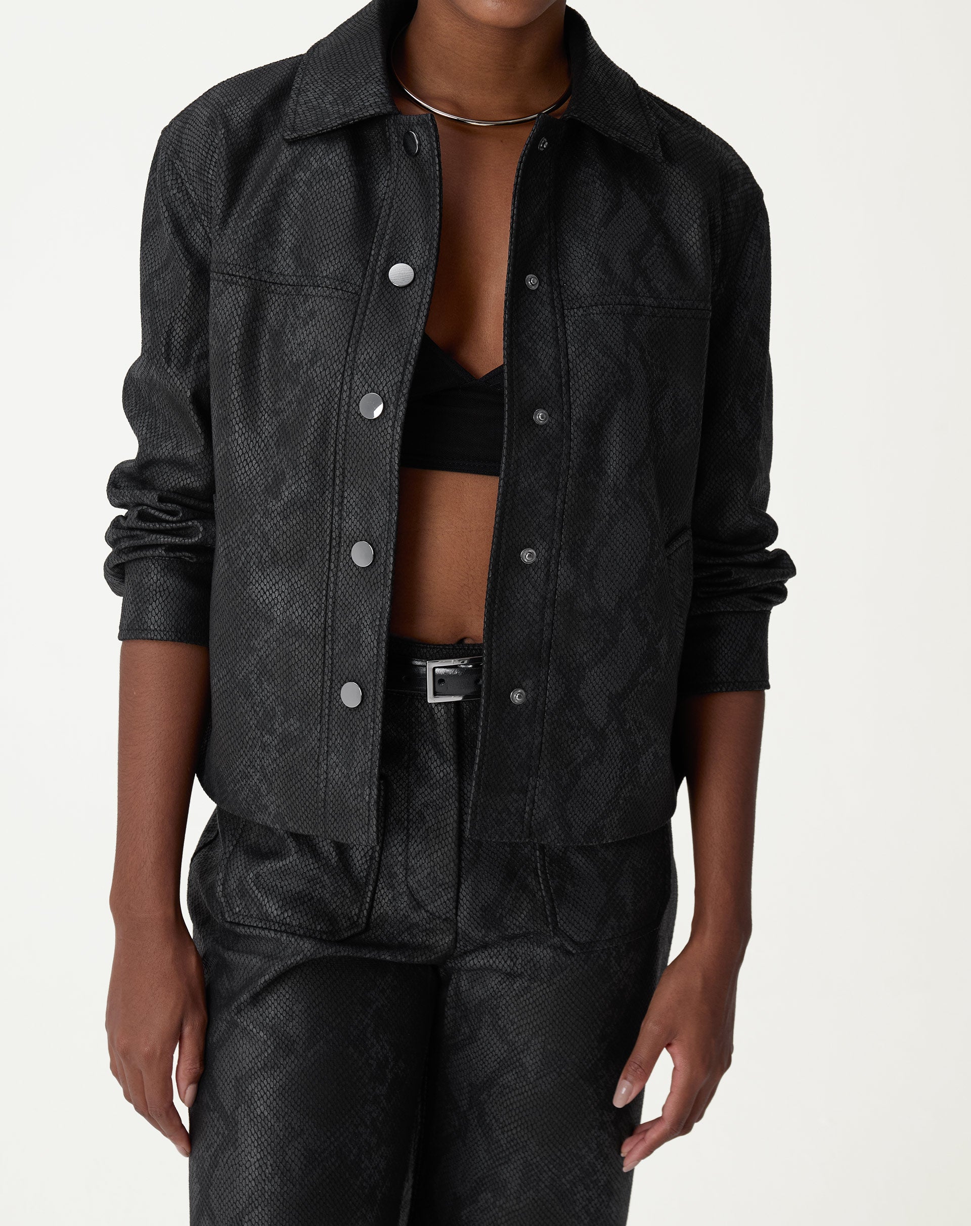 Modelo vestindo jaqueta midi preta em PU estilo cobra, com calça combinando. Apresentação frontal com fechamento aberto.