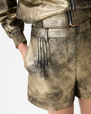 Close nas pregas frontais e bolso lateral do shorts metálico dourado com pespontos visíveis