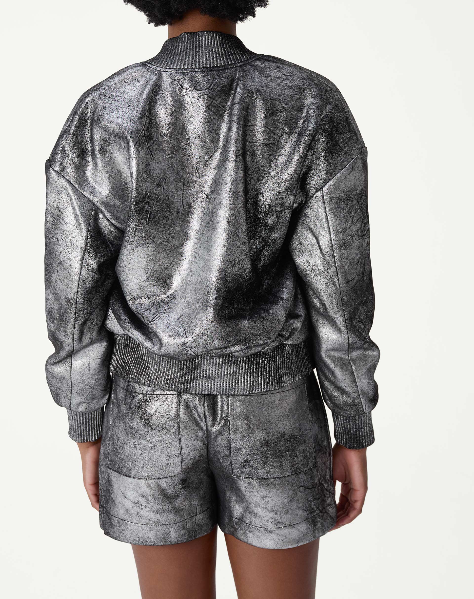 Detalhe em close dos bolsos de aba com botão da jaqueta bomber prata, textura metalizada desgastada e costuras visíveis