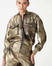 Jaqueta bomber dourada em suedine, modelo vestindo look coordenado com shorts metálicos, vista frontal mostrando zíper e gola canelada