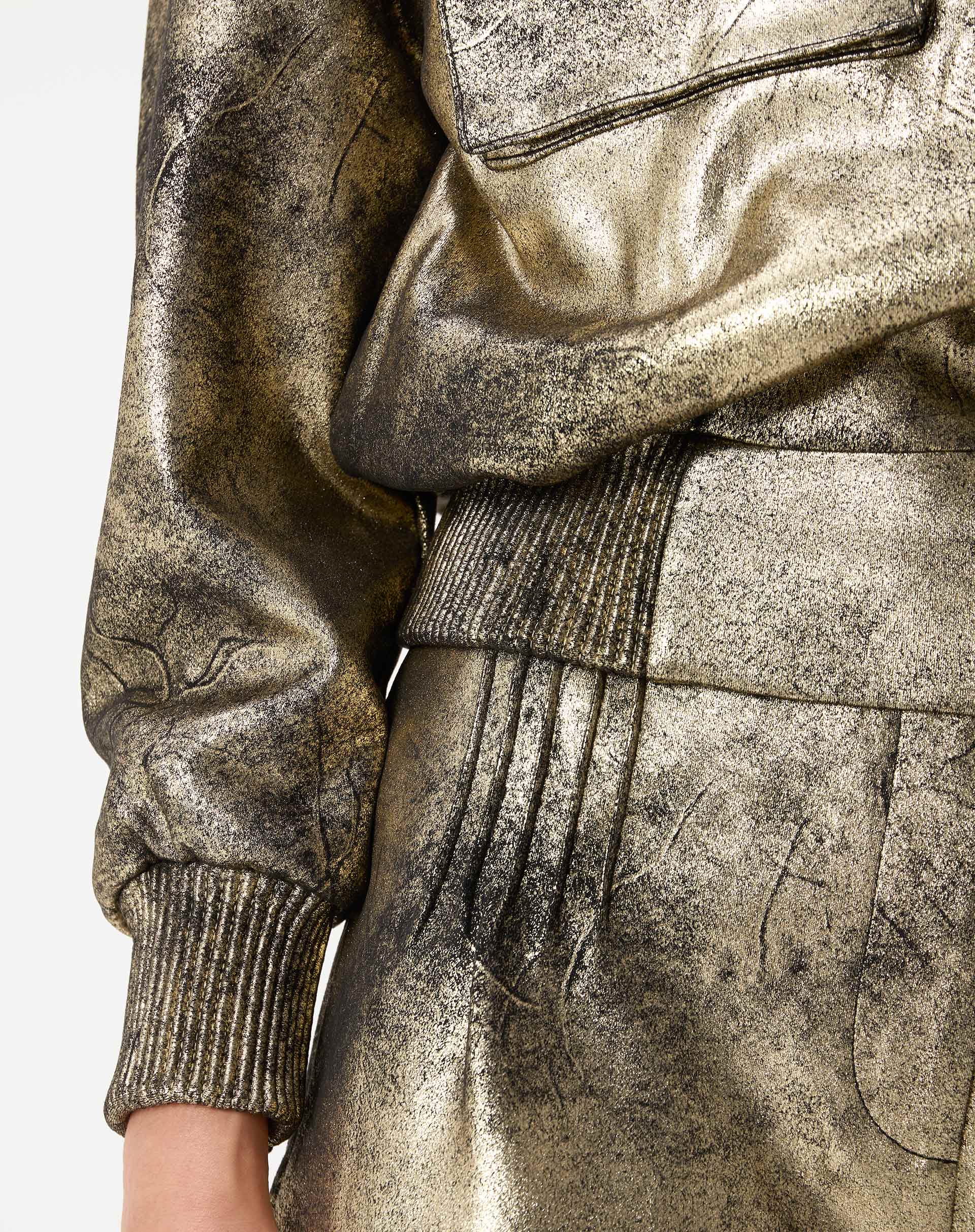 Detalhe da barra e punho canelado da jaqueta bomber dourada e textura do tecido suedine metalizado