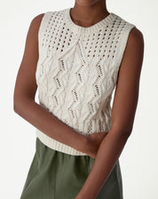 Modelo vestindo regata de tricô bege com ponto diferenciado, detalhamento frontal do torso mostrando cava sem mangas e barra canelada.