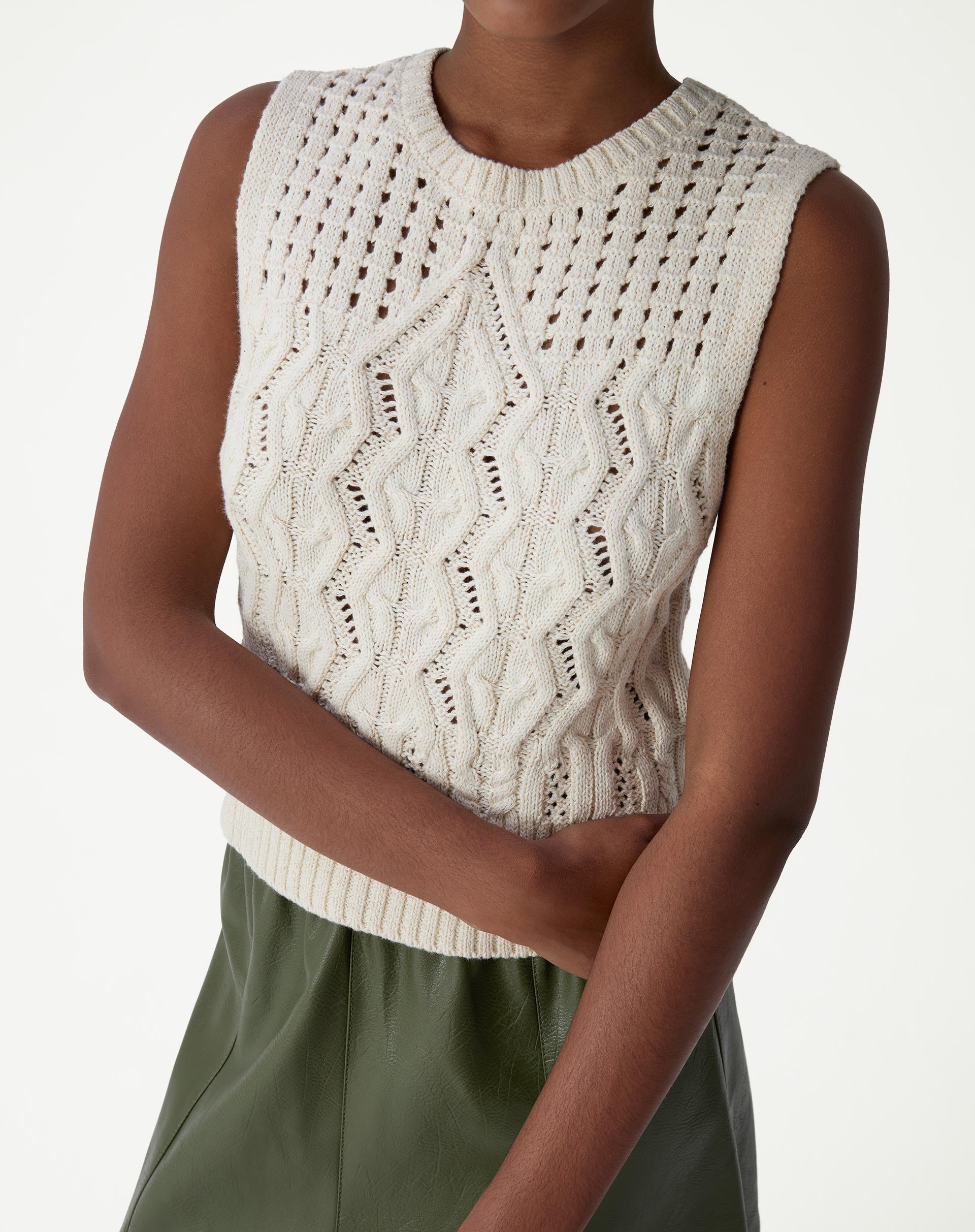 Modelo vestindo regata de tricô bege com ponto diferenciado, detalhamento frontal do torso mostrando cava sem mangas e barra canelada.