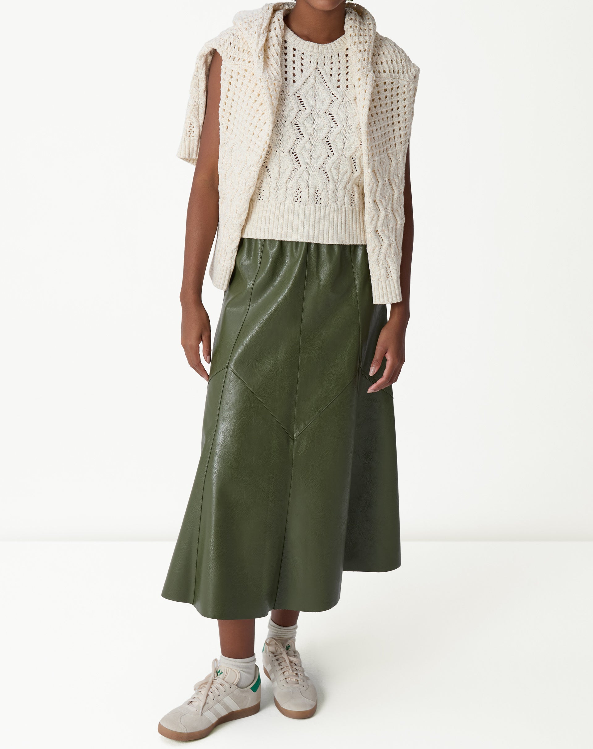 Regata de tricô bege vista frontal com suéter do mesmo tricô drapeado sobre os ombros, destaque para os pontos abertos e tranças.