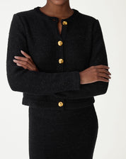 Cardigan preto tricot slim com botões dourados, vista frontal com modelo de braços cruzados e saia midi combinando