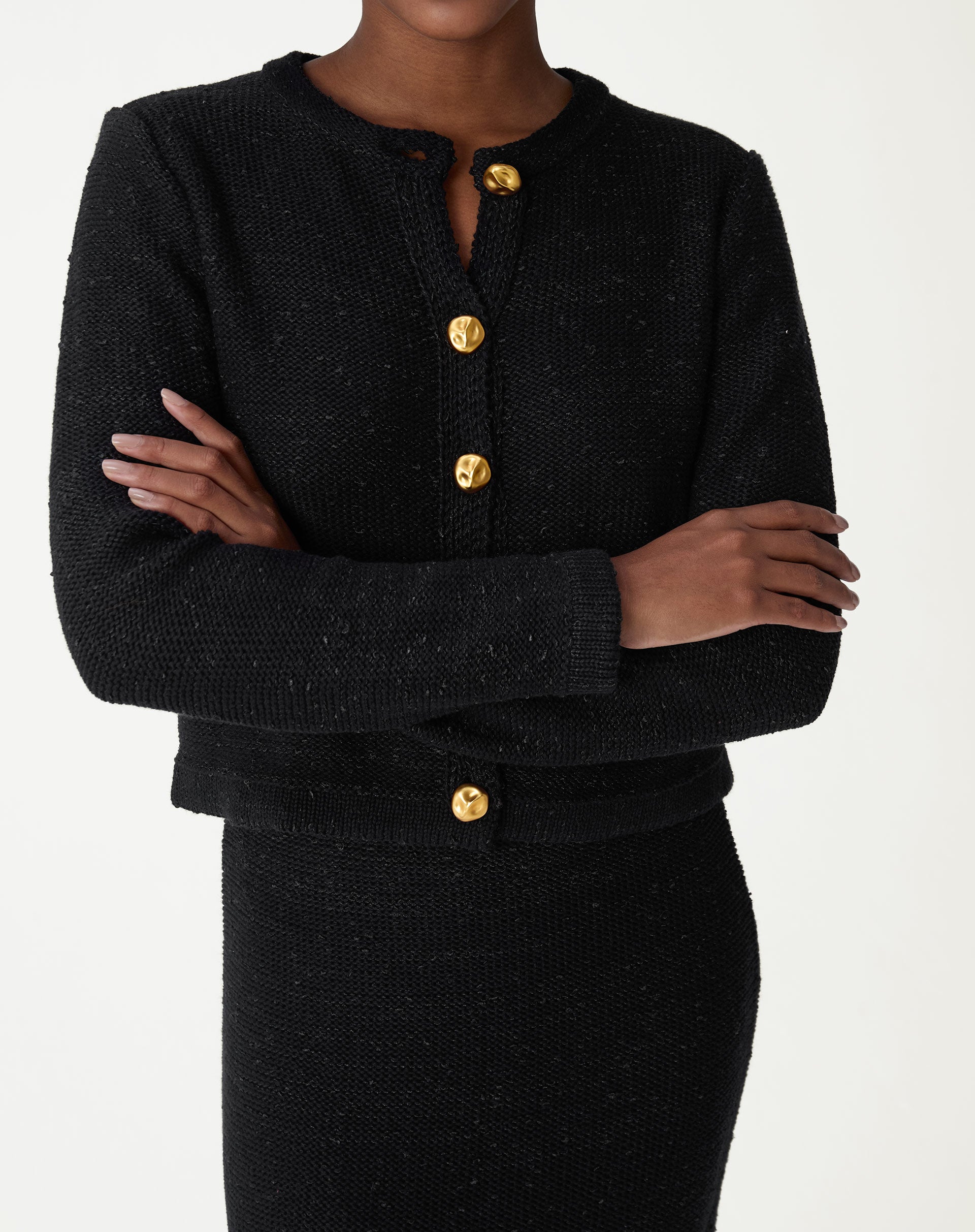 Cardigan preto tricot slim com botões dourados, vista frontal com modelo de braços cruzados e saia midi combinando