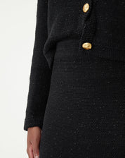 Close do cós alto em tricot preto e detalhe do botão dourado do cardigan sobreposto, mostrando textura da malha