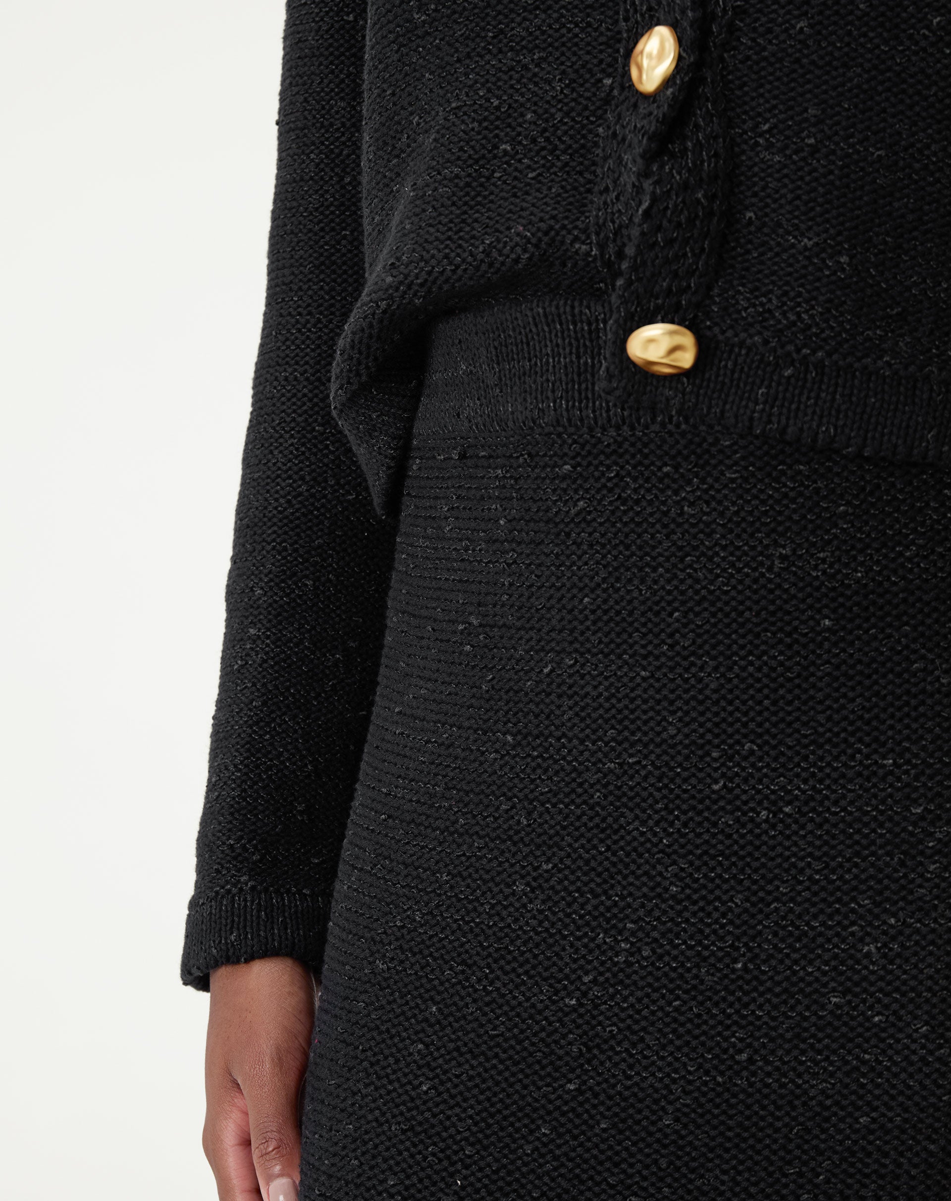 Close do cós alto em tricot preto e detalhe do botão dourado do cardigan sobreposto, mostrando textura da malha
