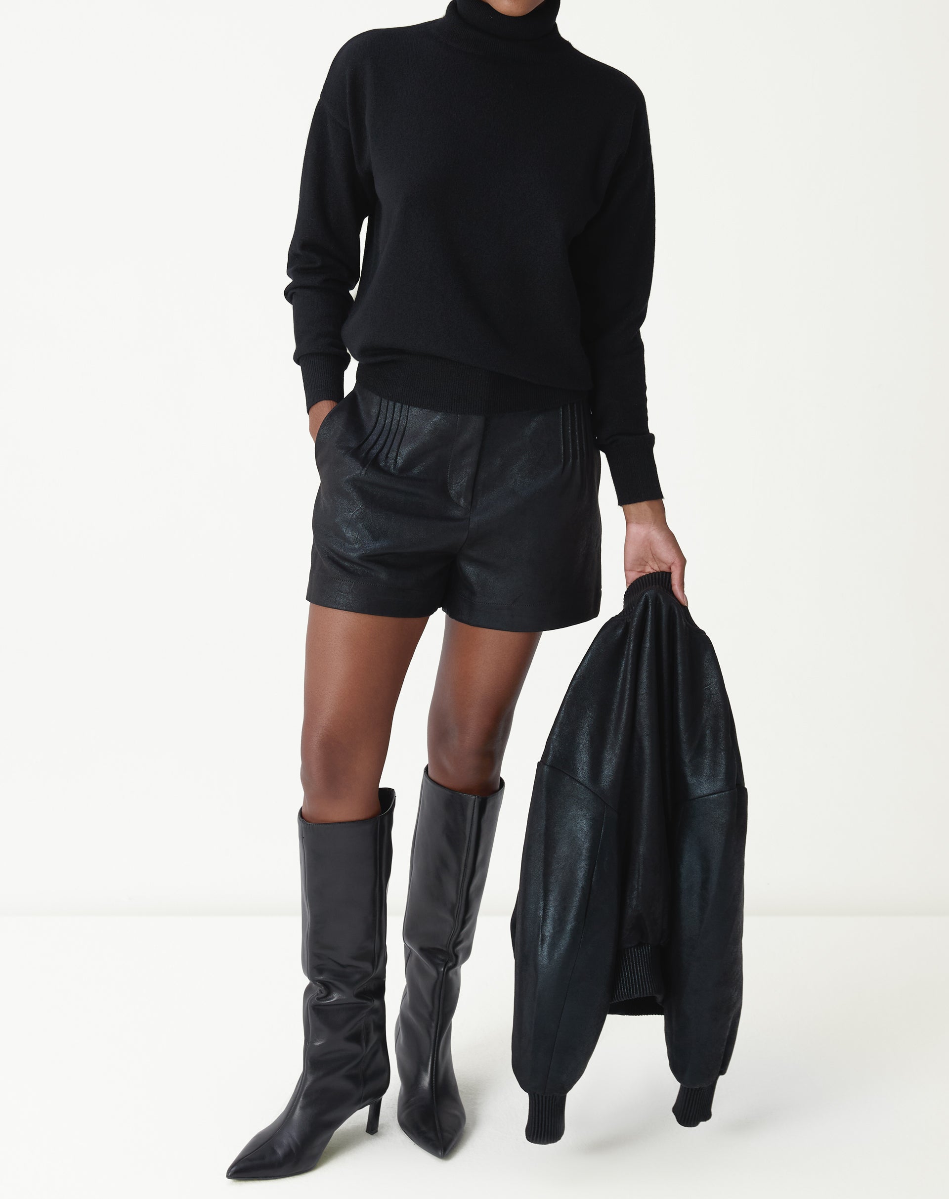Modelo usando suéter gola alta em cashmere preto com shorts de couro e botas longas, look urbano e minimalista