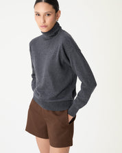 Modelo com suéter cashmere de gola alta em cinza mescla escuro visto de lateral com shorts marrons, estilo casual-chique com fundo branco.