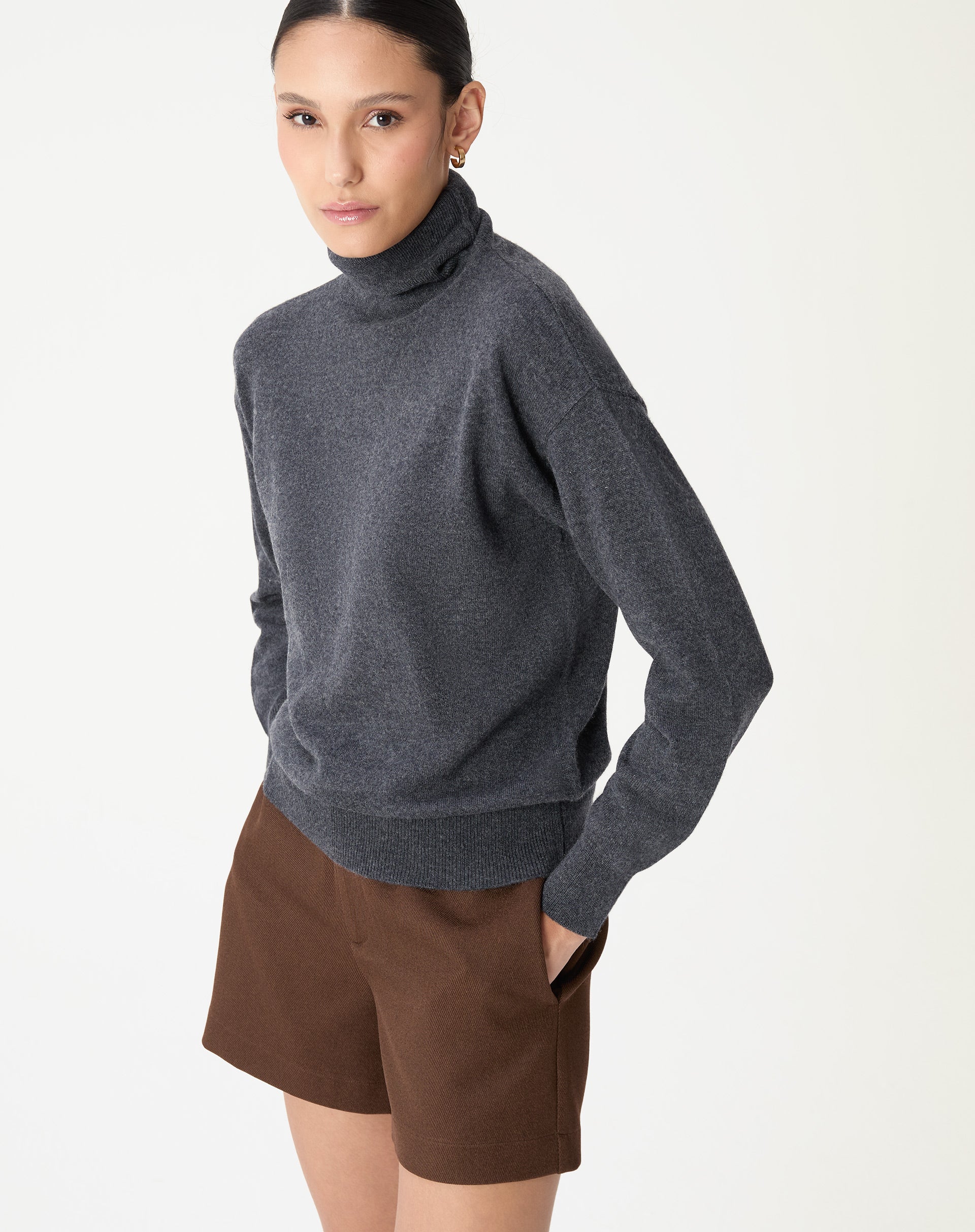 Modelo com suéter cashmere de gola alta em cinza mescla escuro visto de lateral com shorts marrons, estilo casual-chique com fundo branco.