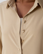 Detalhe lateral mostrando a fenda discreta e a barra arredondada da camisa bege, com punho simples e caimento fluido