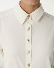 Close-up do colarinho pontudo e do fecho frontal da camisa alongada off-white com botões dourados decorativos, detalhe do tecido fluido