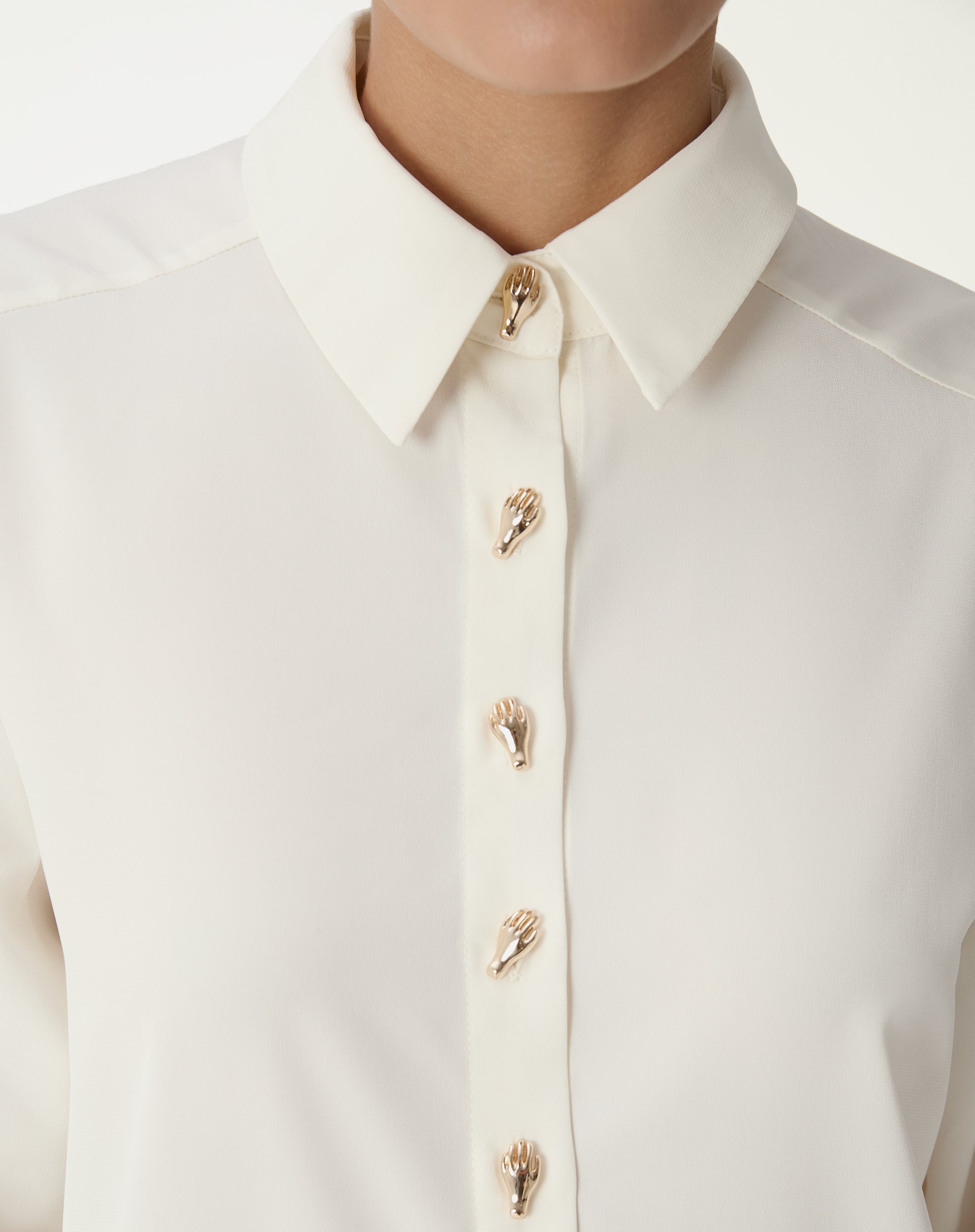 Close-up do colarinho pontudo e do fecho frontal da camisa alongada off-white com botões dourados decorativos, detalhe do tecido fluido