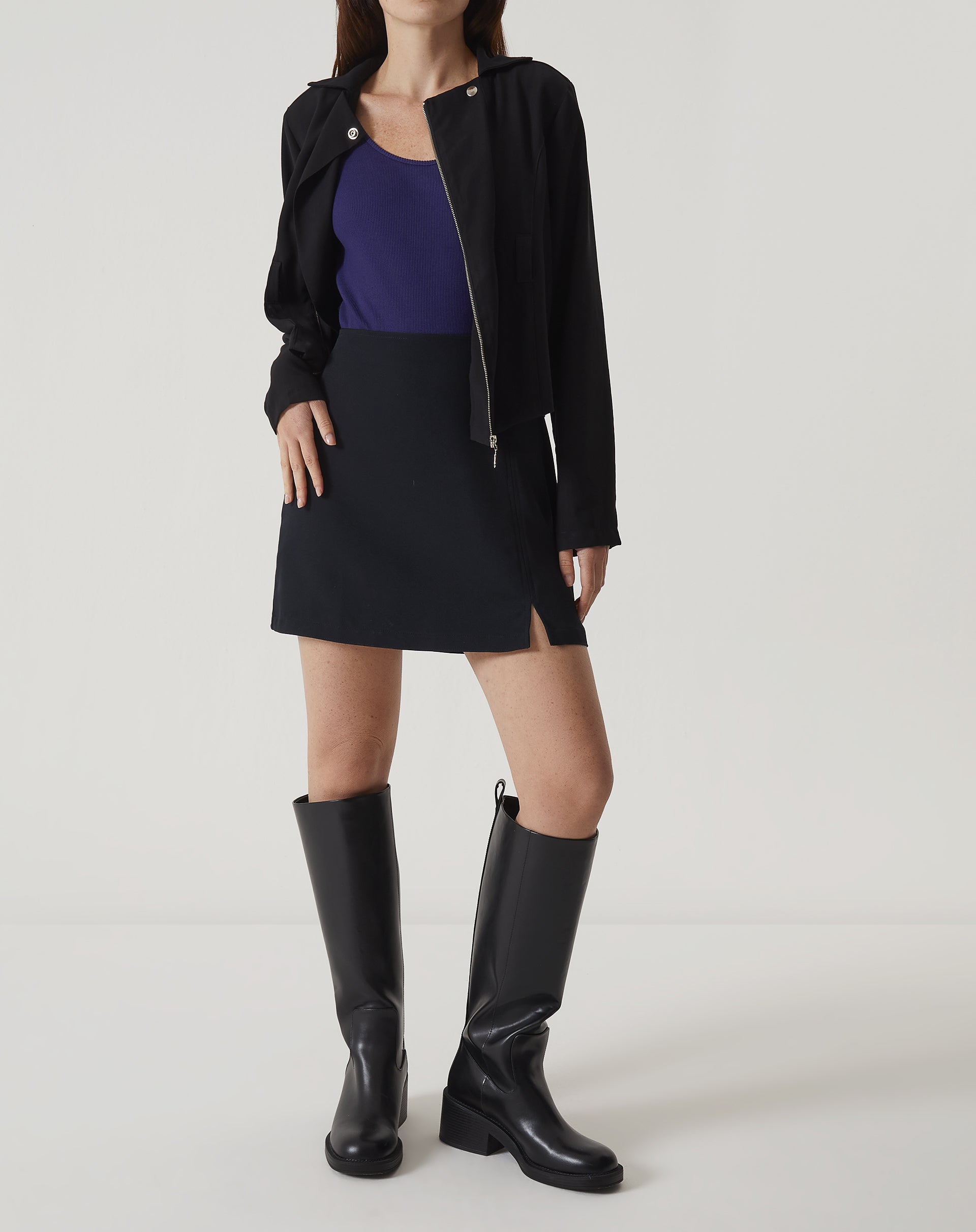Modelo vestindo saia curta de alfaiataria preta com botas e blusa roxa.