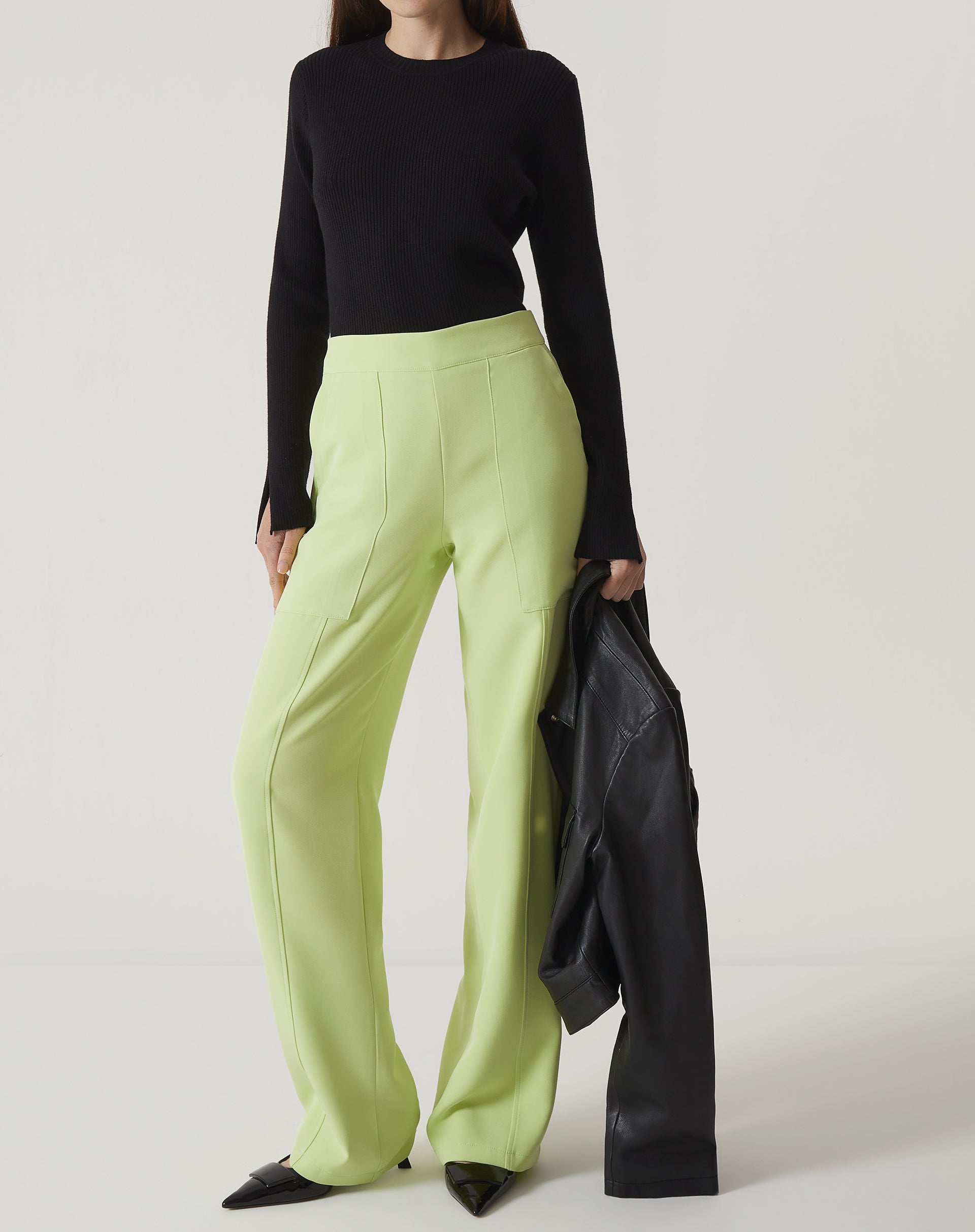 Modelo vestindo calça reta de alfaiataria verde claro com blusa preta e segurando um casaco preto.