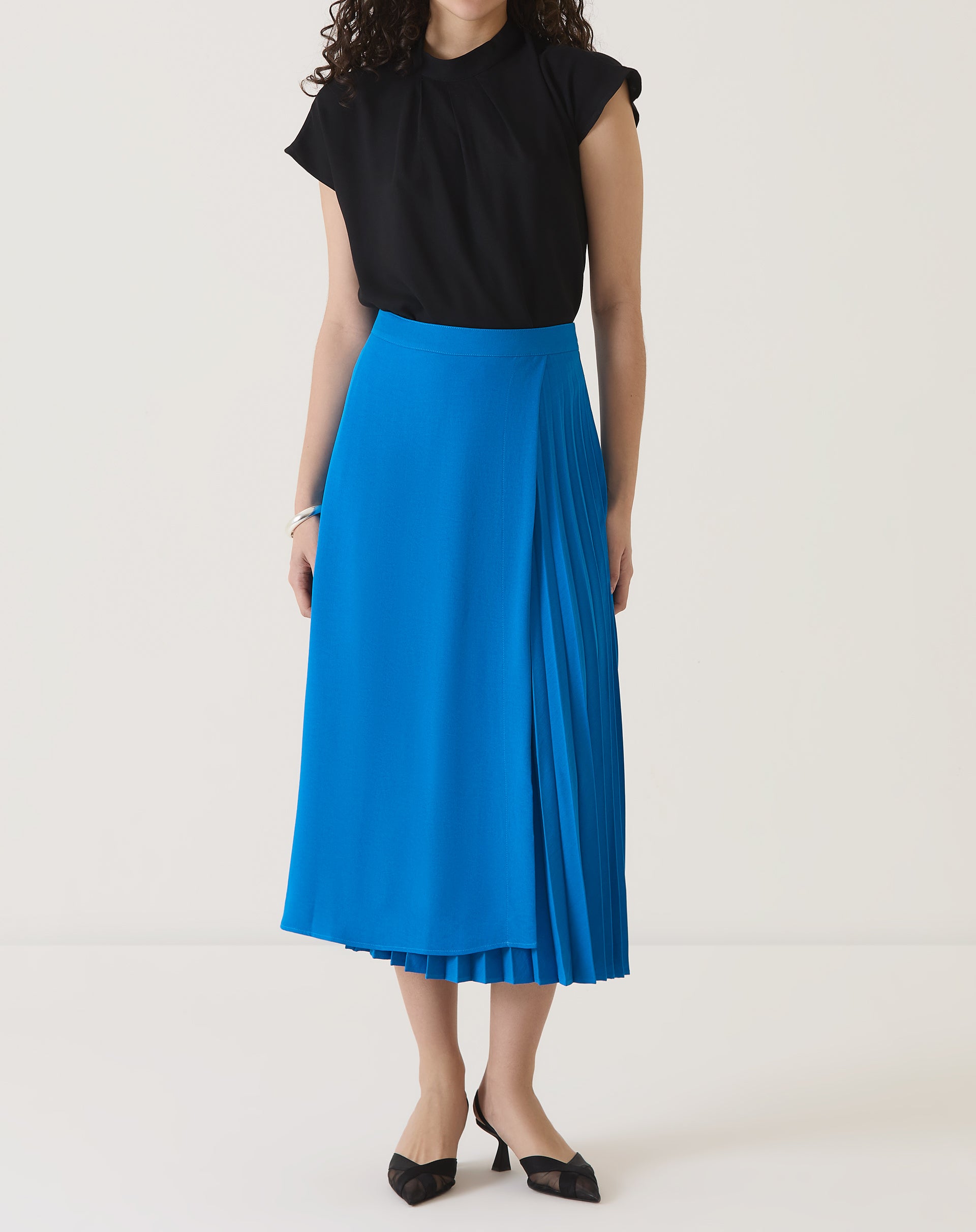 Modelo vestindo saia azul com pregas e blusa preta, vista traseira.