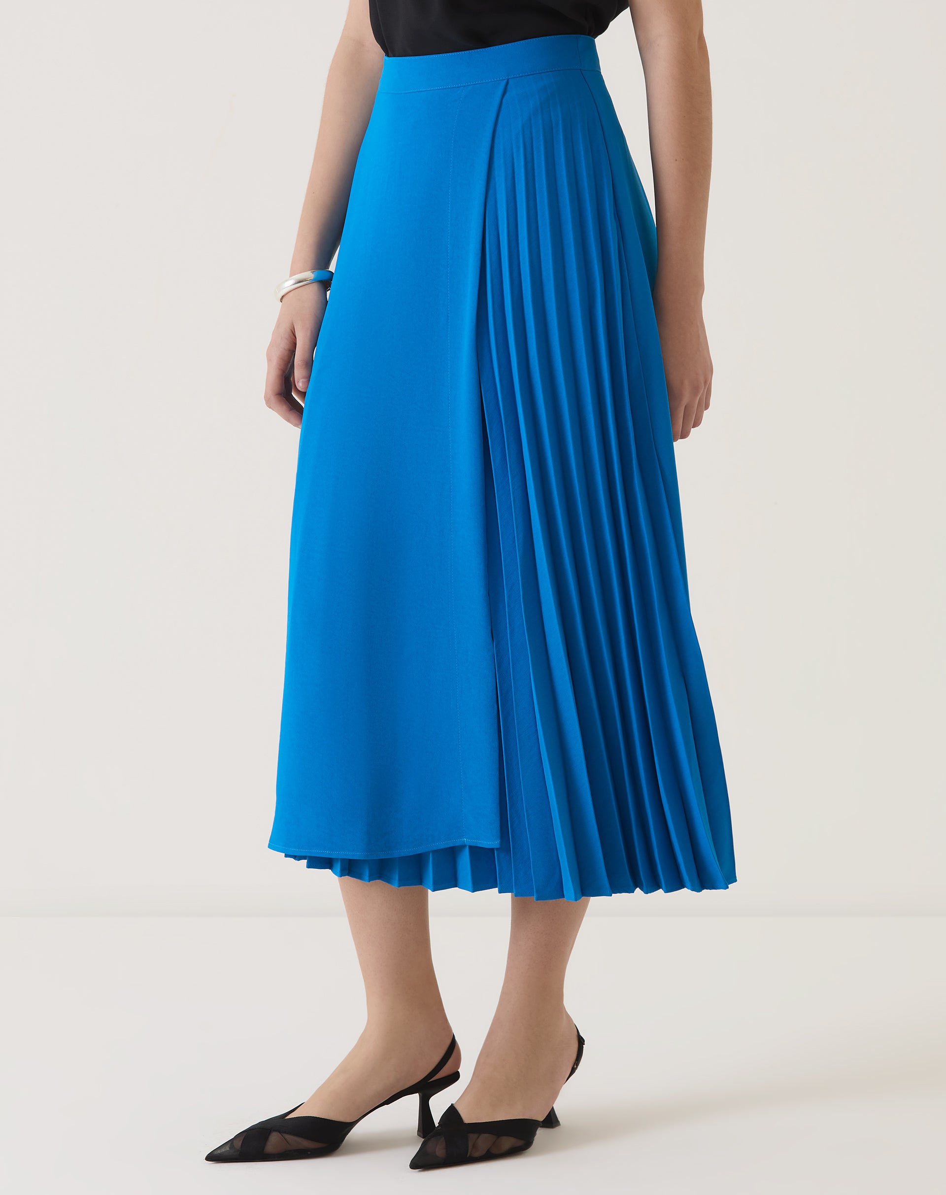 Modelo vestindo saia azul com pregas e blusa preta, vista lateral.