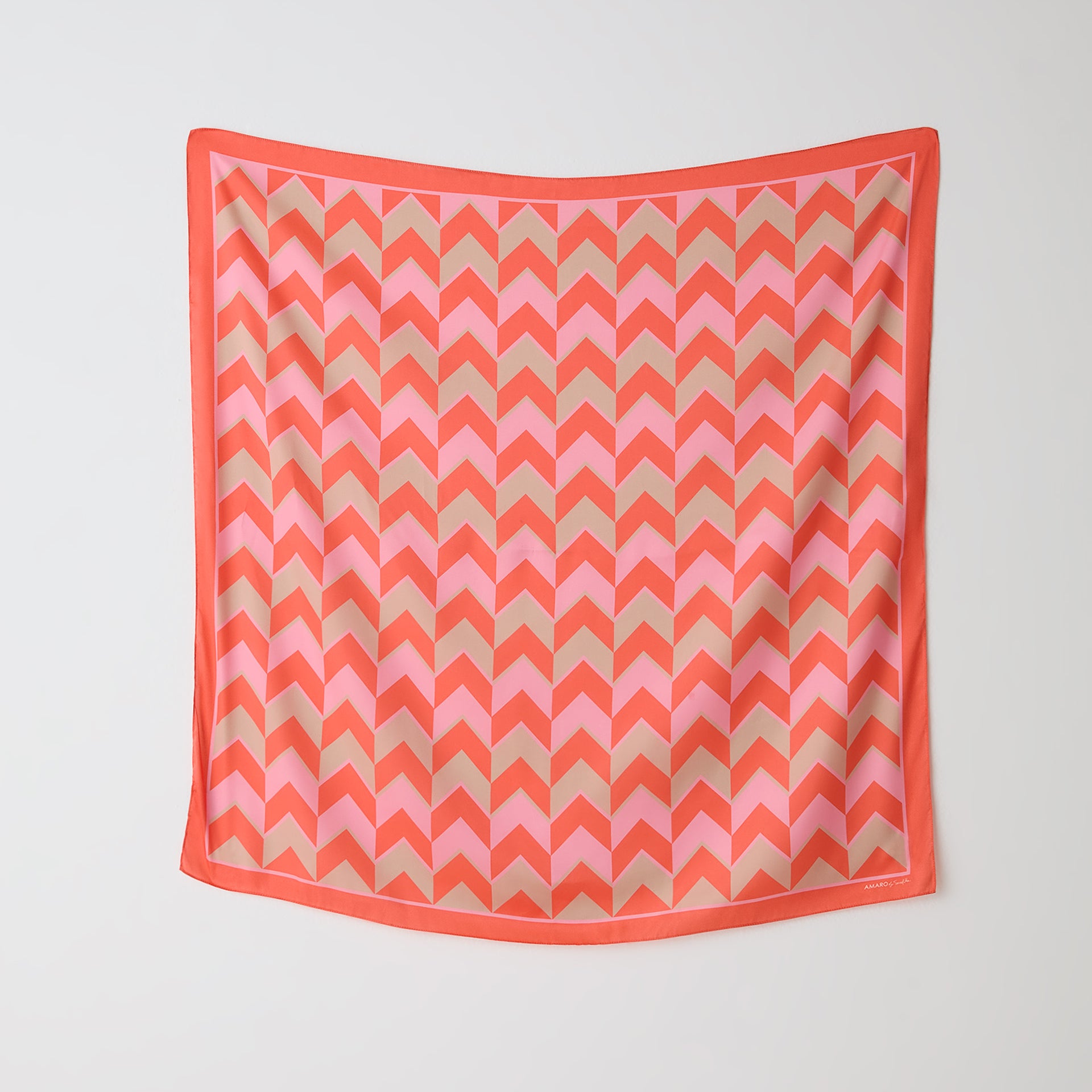 Lenço quadrado AMARO by Scarf Me 90x90cm exibido estendido na parede com estampa chevron coral, rosa e bege, mostrando padrão geométrico e borda coral.