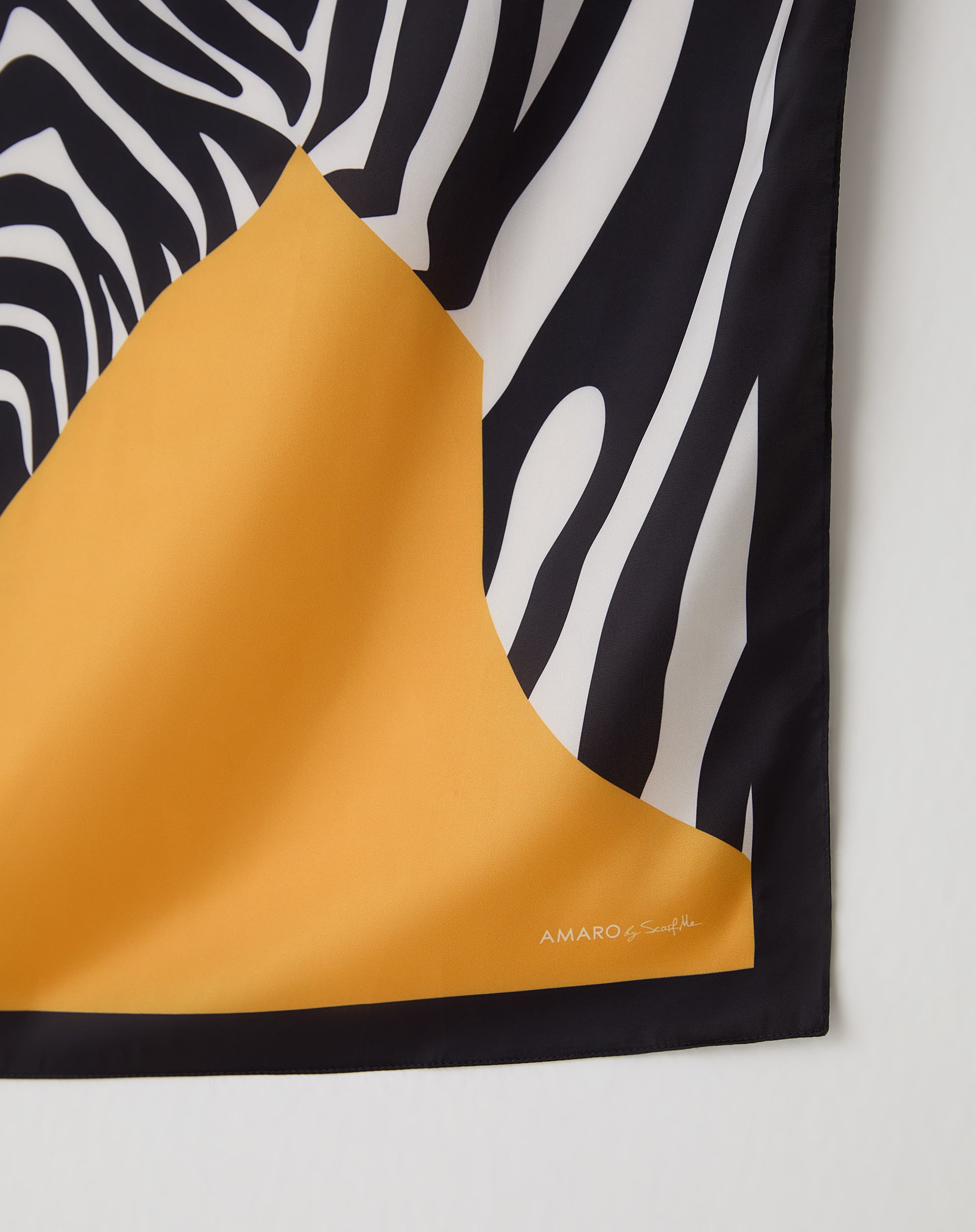Close-up do canto do lenço mostrando barrado preto, assinatura AMARO e contraste entre a estampa zebra e o campo amarelo mostarda